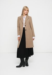 Cappotto in misto lana beige con un design su misura, due bottoni neri, tasche laterali e una texture liscia, indossato sopra una gonna nera plissettata.