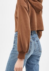 Persona vistiendo una sudadera corta marrón y unos vaqueros de tiro alto azules, mostrada de lado desde la cintura hacia abajo.