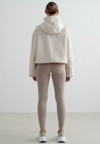 Veste courte beige clair avec capuche, associée à un legging beige ajusté et des chaussures de sport blanches, placée sur un fond neutre.