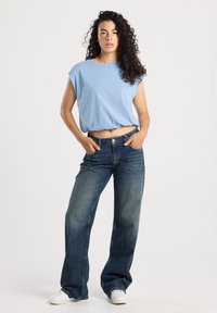 Giovane donna con capelli ricci che indossa una maglietta senza maniche azzurra chiara, pantaloni jeans larghi blu scuro e sneakers bianche, in piedi con le mani nelle tasche.
