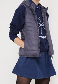 Grå pufferväst med huva, med en kviltad textur, dragkedjestängning och sidofickor, bärs över en marinblå tröja och en denimkjol.