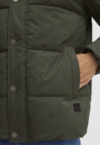 Dark green puffer jacket with a textured fabric, snap buttons, front pocket, and a branded patch on the lower side.