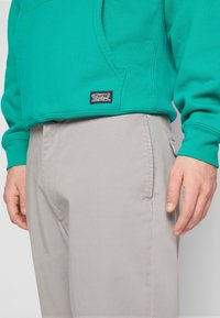 Zelená mikina se klokaní kapsou a štítkem, kombinovaná se světle šedými kalhotami s hladkým povrchem a postranní kapsou.