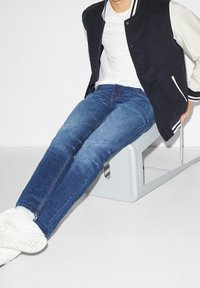 Person som bär en svart och vit varsityjacka, vit skjorta, blå jeans och vita sneakers, sitter på en modern grå stol.