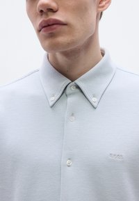 Svetlomodrá polo košeľa s textúrovaným vzorom, dvoma prednými gombíkmi a malým vyšívaným logom na ľavej strane hrudníka. Klasický golier.