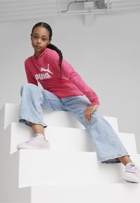 Roza dukserica s bijelim PUMA logotipom, svijetloplave široke traperice i svijetloplave tenisice. Model sjedi na bijelim stepenicama.