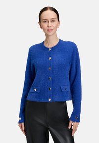 Vrouw in een felblauwe, zachte knopencardigan met twee voorzakken en zwarte leren broek, staand tegen een effen achtergrond.