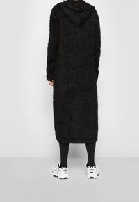 Lång svart fluffig kappa med huva, med långa ärmar och fåll som når nedanför knäna, bärs över svarta leggings och vita sneakers.