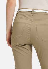 Beige skinny broek met een gladde textuur, voorzien van twee achterzakken en een witte riem. De pasvorm is getailleerd en benadrukt de vorm.
