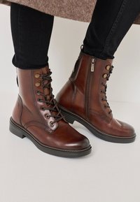 Stivali alla caviglia in pelle marrone con stringhe frontali e cerniera laterale. Presentano occhiali in metallo e una finitura liscia, con un tacco basso a blocco.