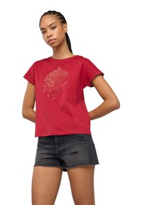 Maglietta rossa in cotone con maniche corte, caratterizzata da bordi ondulati e un design di loto dorato al centro. Abbinata a pantaloni corti neri strappati.