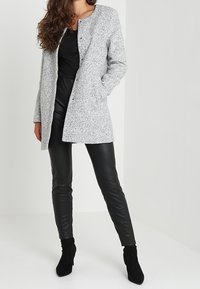 Cappotto grigio maculato con scollatura rotonda e chiusura frontale con bottoni, abbinato a pantaloni in pelle neri e stivaletti alla caviglia. Tessuti lisci in tutto.