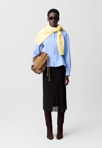 Camicia a righe blu con un maglione giallo poggiato sulle spalle, gonna plissettata nera, stivali alti in suede marrone e una borsa a secchiello marrone.
