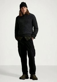 Mörk stickad tröja, grön rutigt skjorta, svarta cargobyxor och mörka skor. Tillbehör inkluderar en svart mössa. Avslappnad, lager-på-lager-look.