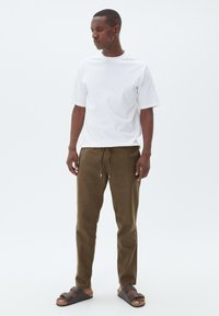 Weißes Kurzarm-T-Shirt, braune Jogginghose mit Kordelzug und braune Sandalen mit Riemen. Das Modell steht vor einem schlichten weißen Hintergrund.