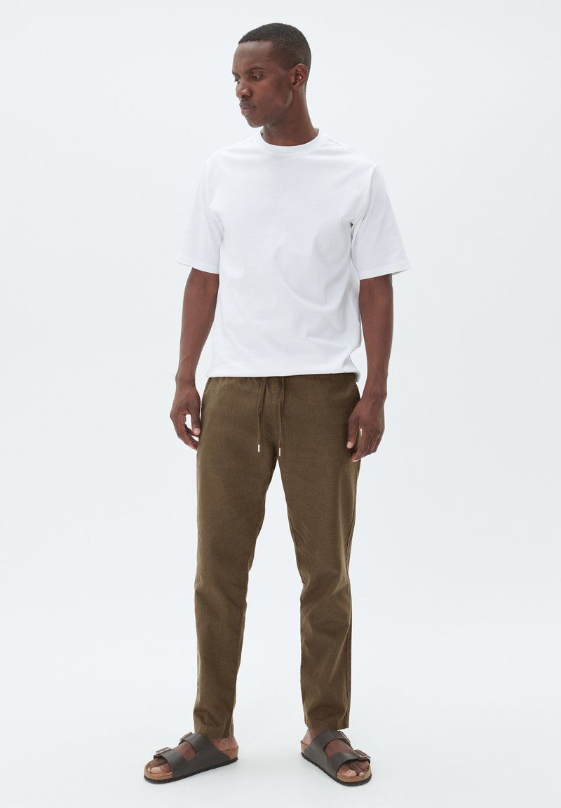 Weißes Kurzarm-T-Shirt, braune Jogginghose mit Kordelzug und braune Sandalen mit Riemen. Das Modell steht vor einem schlichten weißen Hintergrund.