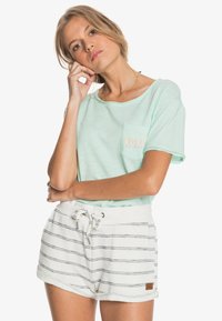 Pistaziengrünes Baumwoll-T-Shirt mit einer kleinen Tasche und dem "ROXY"-Logo. Kombiniert mit weißen gestreiften Shorts mit Kordelzug und strukturiertem Finish.
