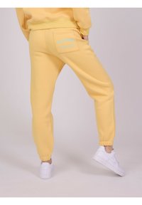 Gule sweatpants lavet af blødt stof med elastisk talje, ribbede ankler og en baglomme med lyseblå tekst.
