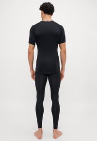 Un bărbat stând descult, cu spatele, îmbrăcat într-un tricou sport negru cu mânecă scurtă și colanți negri de compresie, pe un fundal simplu.