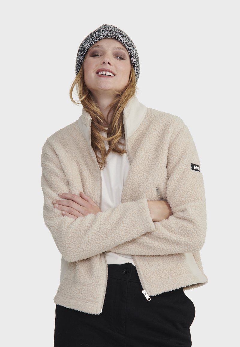 Béžová fleece bunda s vysokým golierom, zipsom a bočnými vreckami. Má kontrastnú krémovú podšívku a čiernu nášivku s logom na rukáve.
