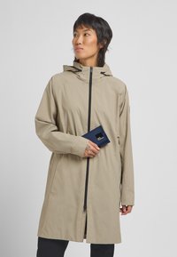 Beige waterproof jacket with a front zipper, adjustable hood, and two side pockets. Model holds a navy wallet featuring a logo.