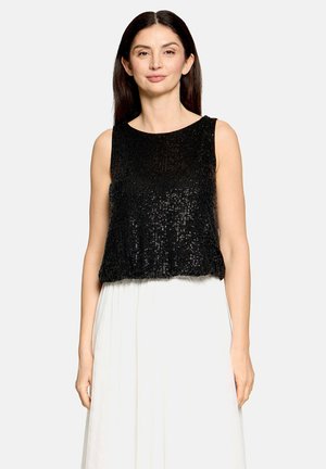 Žena s dlhými tmavými vlasmi, ktorá má na sebe čierny bezrukávový top so sequins a bielu sukňu s vysokým pásom na jednoduchom svetlom pozadí.