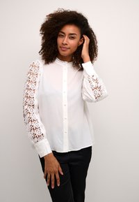 White blouse with lace long sleeves, buttoned front, and a rounded collar. Features floral lace patterns along the sleeves and smooth fabric.