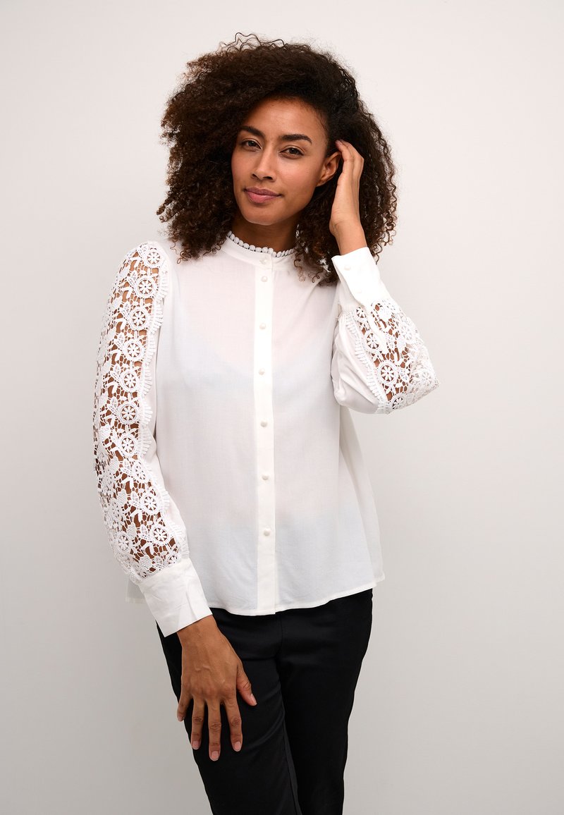 White blouse with lace long sleeves, buttoned front, and a rounded collar. Features floral lace patterns along the sleeves and smooth fabric.