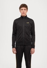 Black tracksuit made of smooth fabric; features a high collar, gold branding, zip closure, and yellow side stripes on pants and jacket.