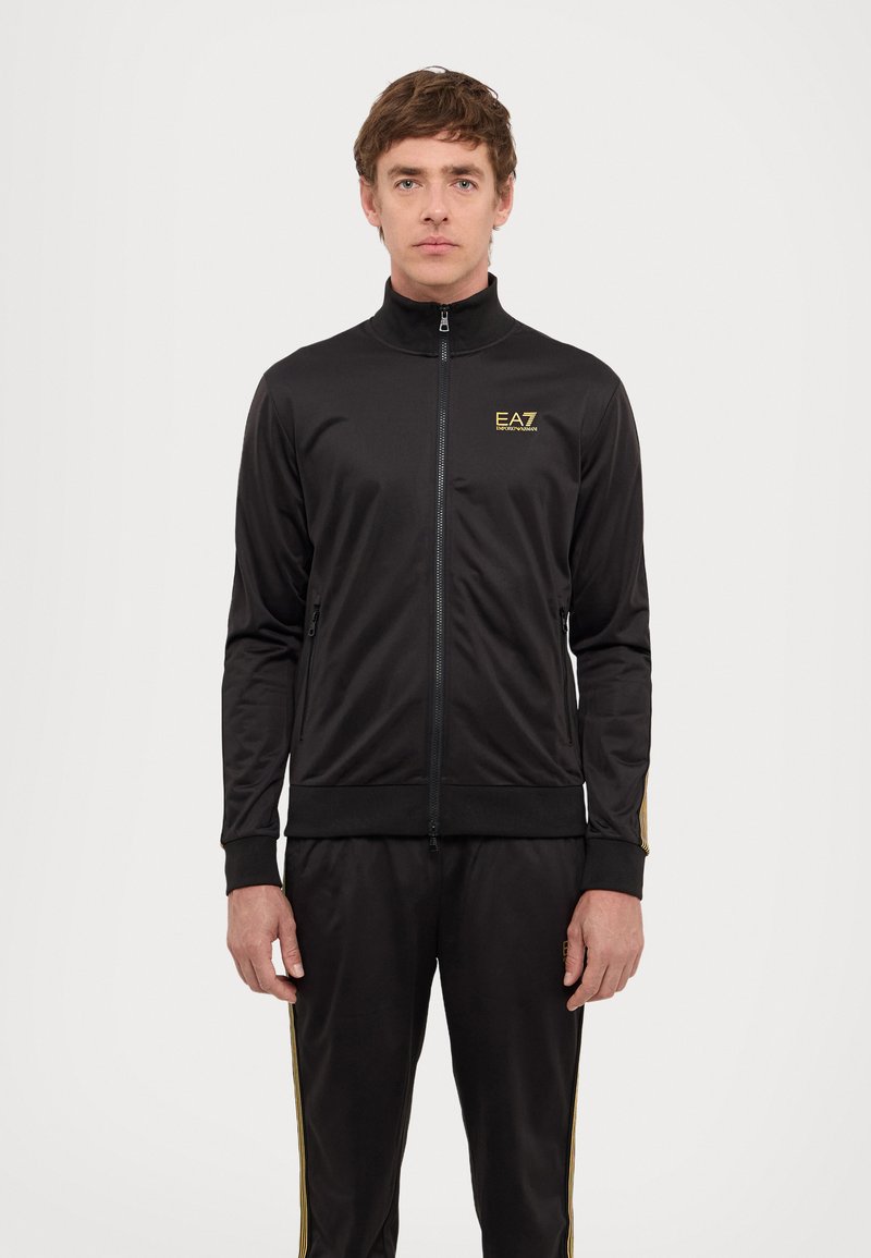 Black tracksuit made of smooth fabric; features a high collar, gold branding, zip closure, and yellow side stripes on pants and jacket.