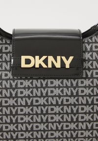 Mustak sintetik handbag dengan flap mengkilap yang menampilkan logo DKNY berwarna emas. Badan tas dihiasi dengan pola DKNY hitam dan putih yang berulang.