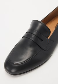 Mocassin en cuir noir avec un bout pointu, présentant un design cousu et une large sangle décorative sur le dessus. L'intérieur présente un rembourrage beige.