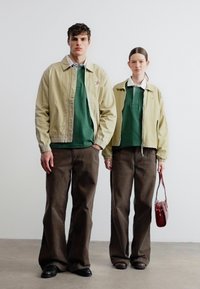 Δύο άτομα στέκονται δίπλα-δίπλα φορόντας μπεζ μπουφάν, πράσινα πόλο πουκάμισα, καφέ παντελόνια και σκούρα παπούτσια· το ένα κρατά κόκκινη τσάντα χειρός.