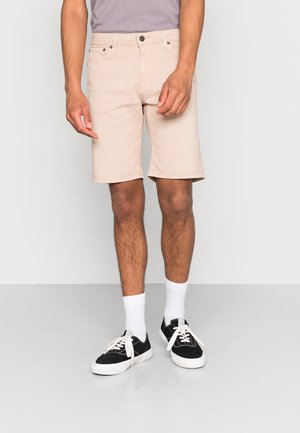 Shorts en denim beige clair à coupe ajustée, avec poches avant et fermeture à bouton. Portés avec des baskets noires et des chaussettes blanches montantes.