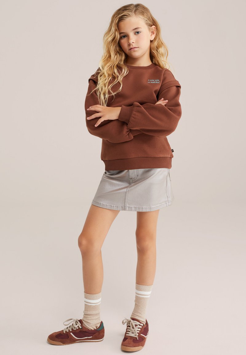 Rjava sweatshirt z nabranimi rokavi, v kombinaciji s srebrno denim krilom. Obut z burgundskimi supergami in svetlo črtastimi nogavicami.