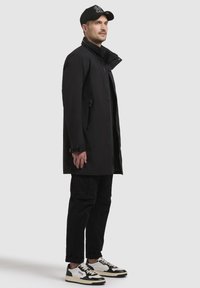 Abrigo negro impermeable con cuello alto, botones frontales, bolsillos laterales con cremallera, combinado con pantalones cargo negros y zapatillas blancas con detalles negros.
