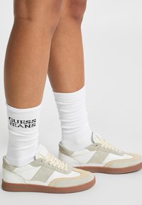 Fehér és bézs sportcipők sima bőrből és vel�úrból készült felsőrésszel, barna gumitalppal. Fehér, bordázott zoknival viselve, amelyen a "GUESS JEANS" felirat látható.