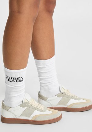 Baskets blanches et beiges avec une tige en cuir lisse et en daim, dotées d'une semelle en caoutchouc beige. Portées avec des chaussettes blanches côtelées affichant "GUESS JEANS".