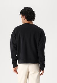 Sudadera negra con cuello redondo acanalado y mangas largas. Presenta un corte relajado y una textura suave. Se lleva sobre pantalones de color claro.