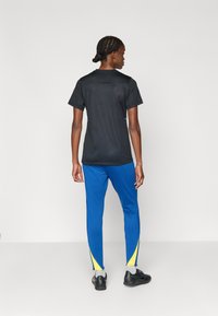 Μαύρο αθλητικό μπλουζάκι κοντομάνικο με στρογγυλή λαιμόκοψη, συνδυασμένο με μπλε αθλητικά παντελόνια με κίτρινες λεπτομέρειες στα πλάγια και μαύρα παπούτσια.