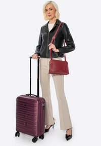 Valigia rigida viola con design rigato e manico retrattile. Giacca di pelle nera, pantaloni beige e borsa a tracolla rossa.