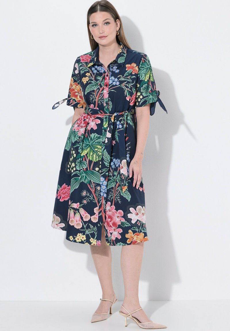 Kobieta w granatowej, kwiecistej sukience midi z krótkimi rękawami, z paskiem w talii i beżowych czółenkach na obcasie typu slingback, stojąca na białym tle.