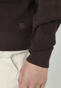 Pull à côtes marron foncé avec une finition texturée, orné d'un petit logo en relief sur l'ourlet, assorti à un pantalon en velours côtelé crème.