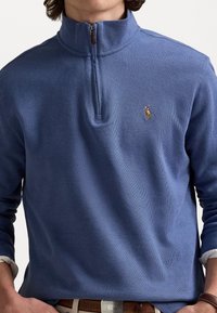 Suéter de punto en azul claro con cuello de media cremallera, textura de punto y logo bordado en el pecho. Mangas largas y ajuste relajado.