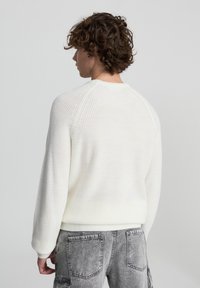 Biały sweter z dzianiny o prążkowanej fakturze, z okrągłym dekoltem i rękawami raglanowymi, w połączeniu z szarymi spodniami dżinsowymi z tylnymi kieszeniami.