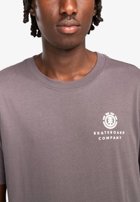 Grijze katoenen t-shirt met korte mouwen, voorzien van een wit logo op de linkerborst en een ronde halslijn. Gladde textuur.