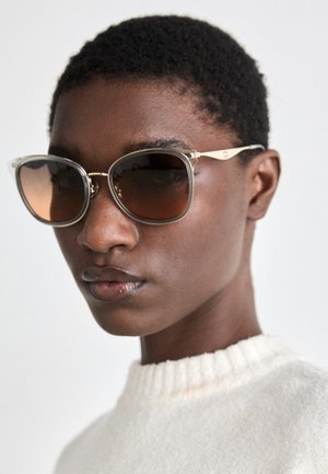 Prevelike kvadratne sončne očala z rjavimi stekli, prozornimi plastičnimi okvirji in zlatimi detajli. Tekstura na površini z minimalističnim dizajnom.