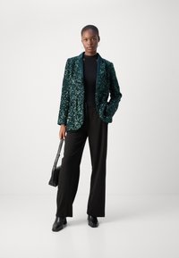 Paljetter blazer i mørkegrønn med klaffekrage, kombinert med svart topp og vide bukser. Modellen holder en liten svart håndveske.