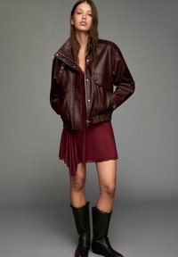 Kvinna iklädd en burgundy läderjacka över en matchande burgundy slipklänning och svarta knähöga läderstövlar, stående mot en grå bakgrund.