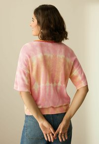 Kurzärmliges Strickoberteil im rosa und gelben Batikmuster, mit geripptem Ausschnitt und Saum. Über hoch taillierten blauen Jeans getragen, von hinten betrachtet.